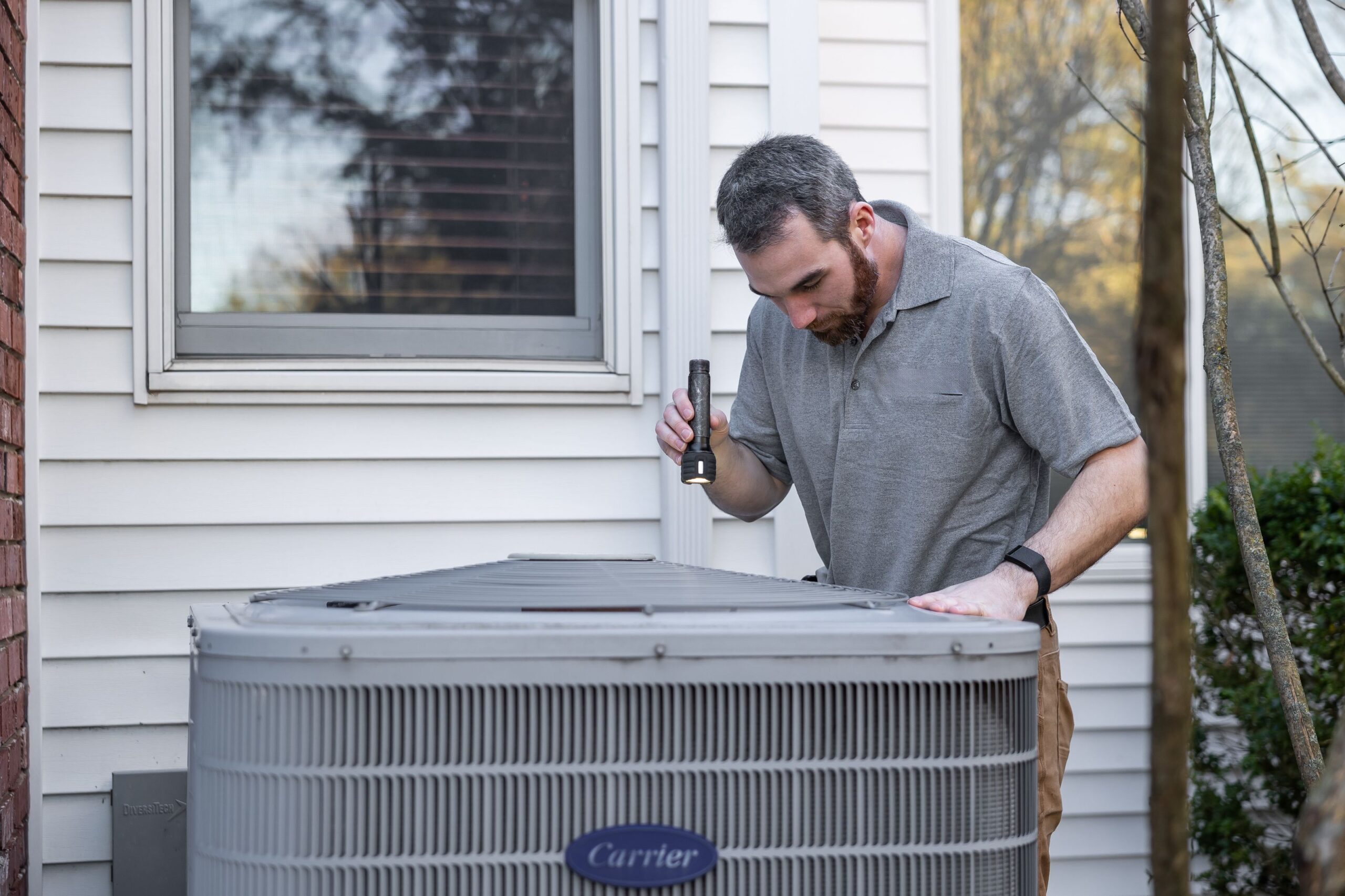HVAC technician inspecting a residential Carrier air conditioning unit with a flashlight