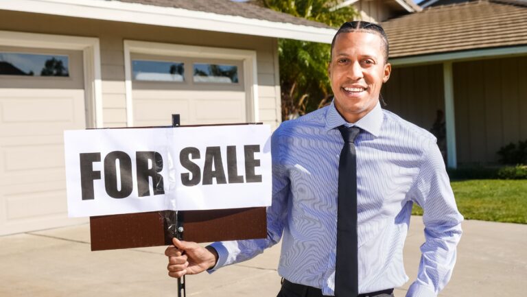 Smiling real estate agent standing beside a for-sale sign in front of a suburban home
