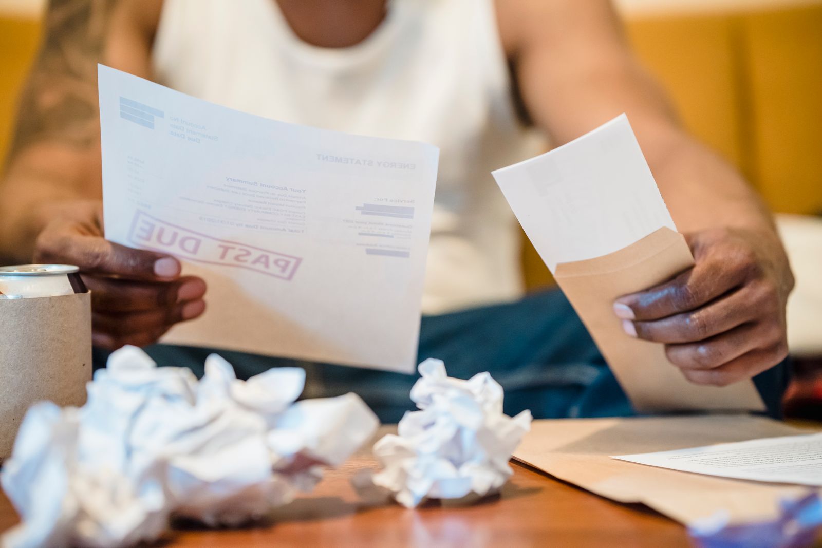 A person at a kitchen table holding a past-due energy bill surrounded by crumpled paperwork