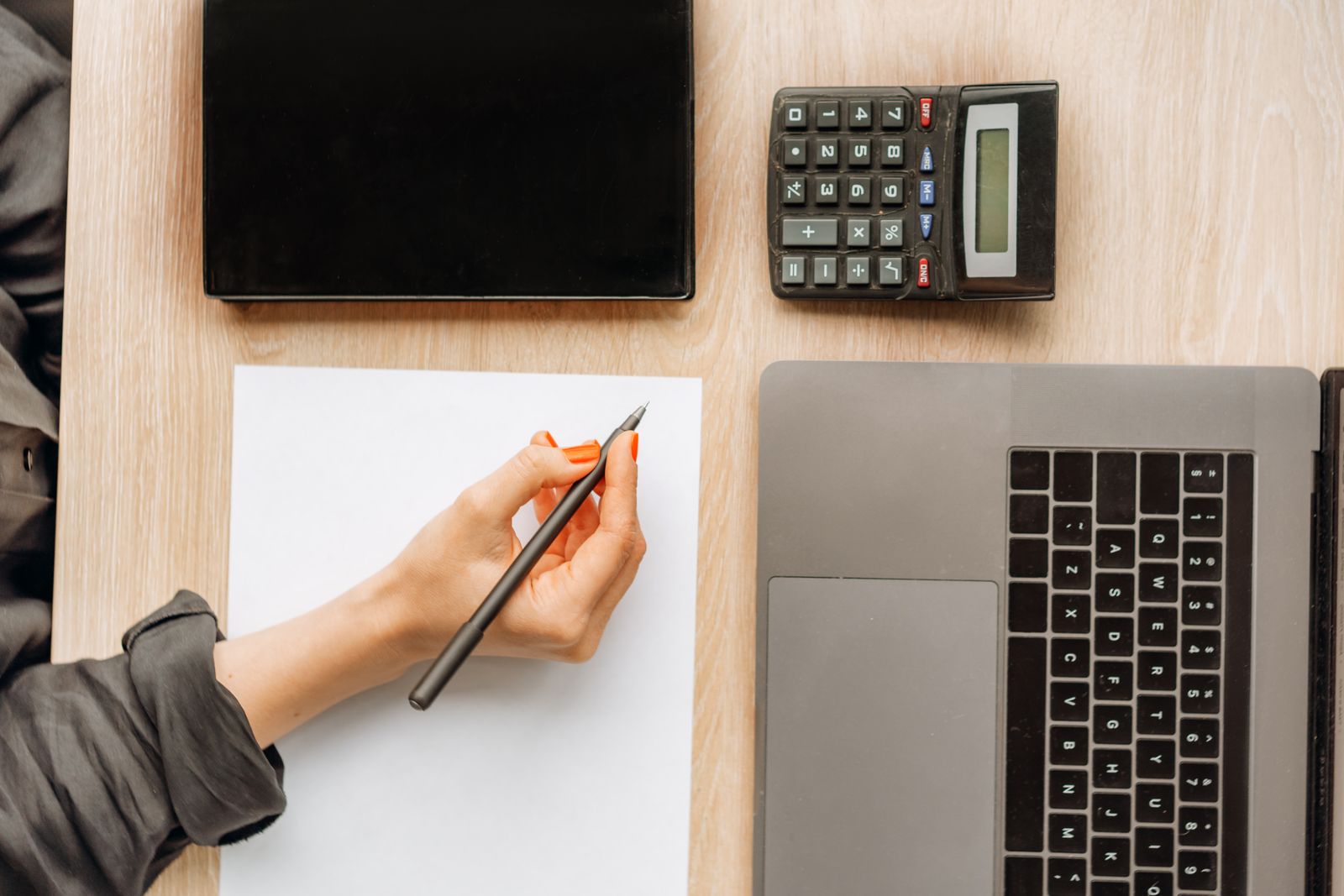 A hand with a pen doing calculations next to a calculator and laptop on a desk