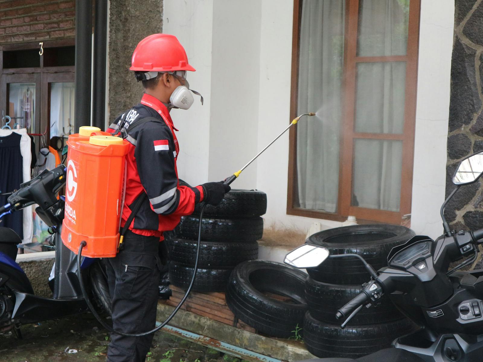Pest control technician in red helmet and respirator using a sprayer to treat the exterior of a residential property