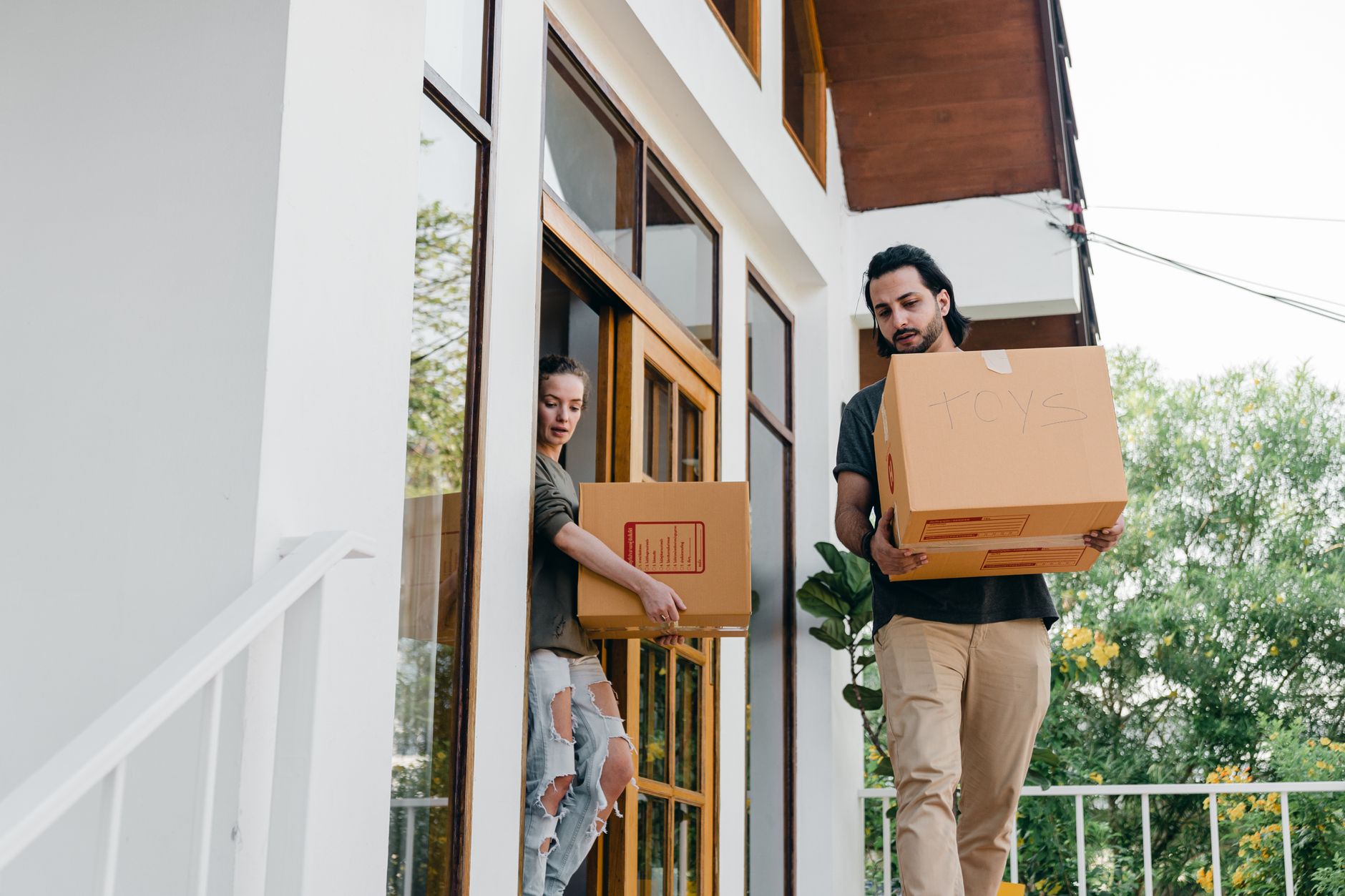 A couple carrying labeled moving boxes onto the porch of their new home on moving day