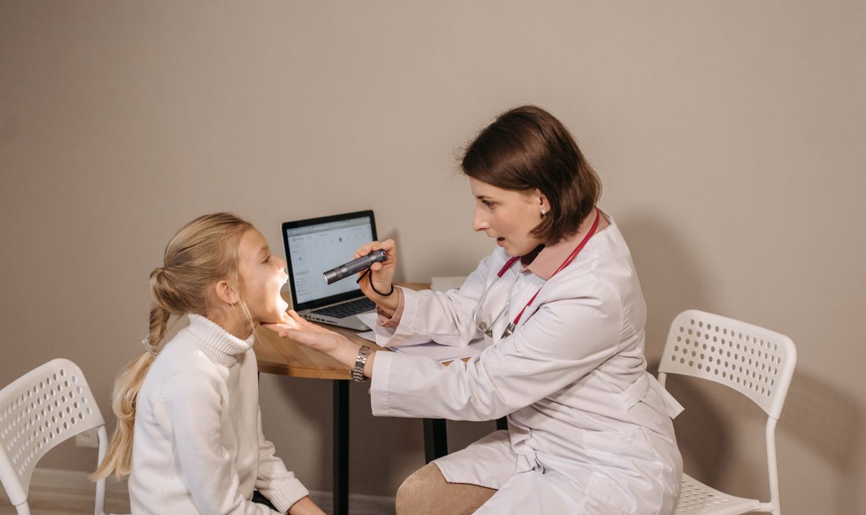 Female pediatrician examining a young child's throat with a flashlight in a clinic setting