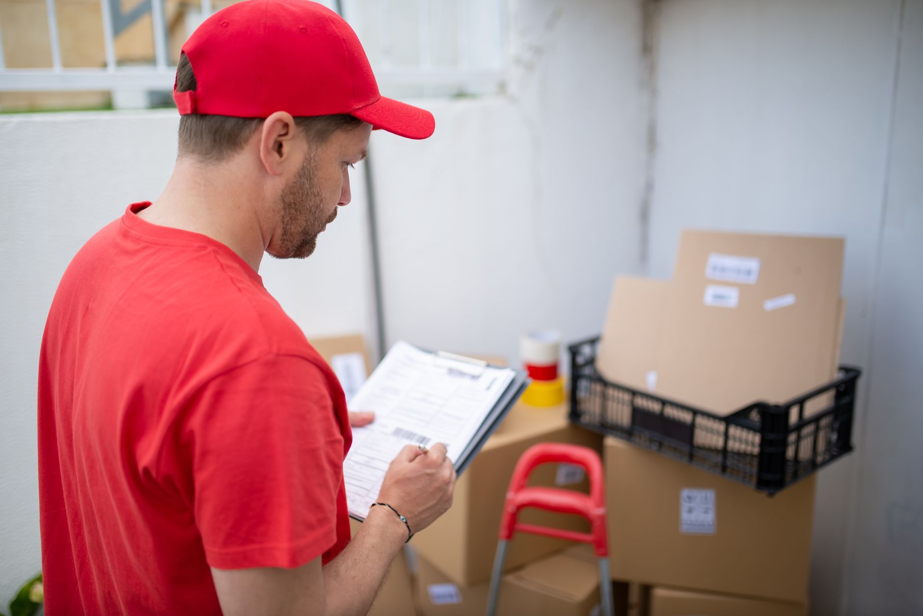 A mover in a red shirt and cap writing up a moving estimate on a clipboard next to packed boxes