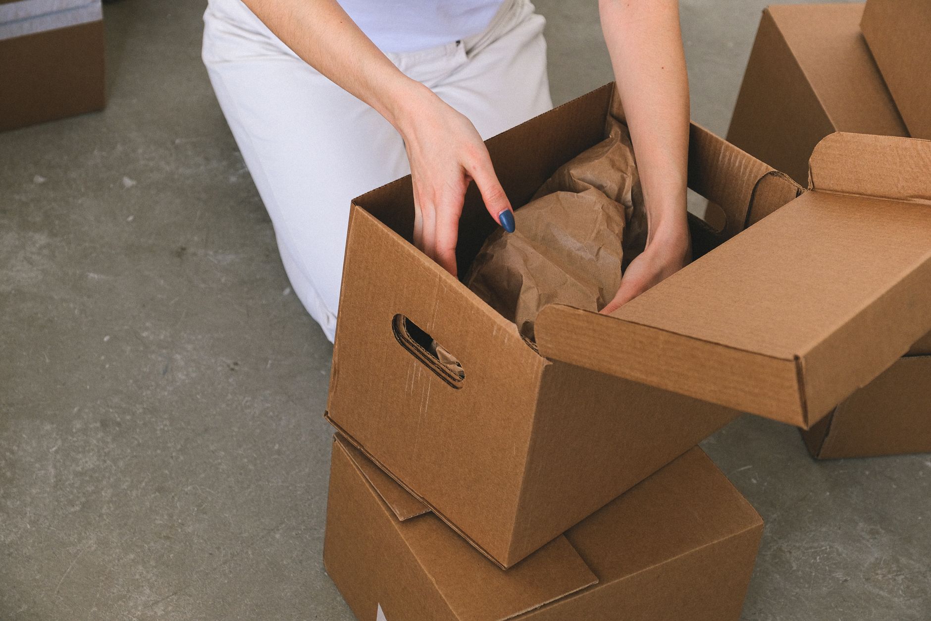 A person kneeling on the floor packing wrapped items into a labeled cardboard moving box