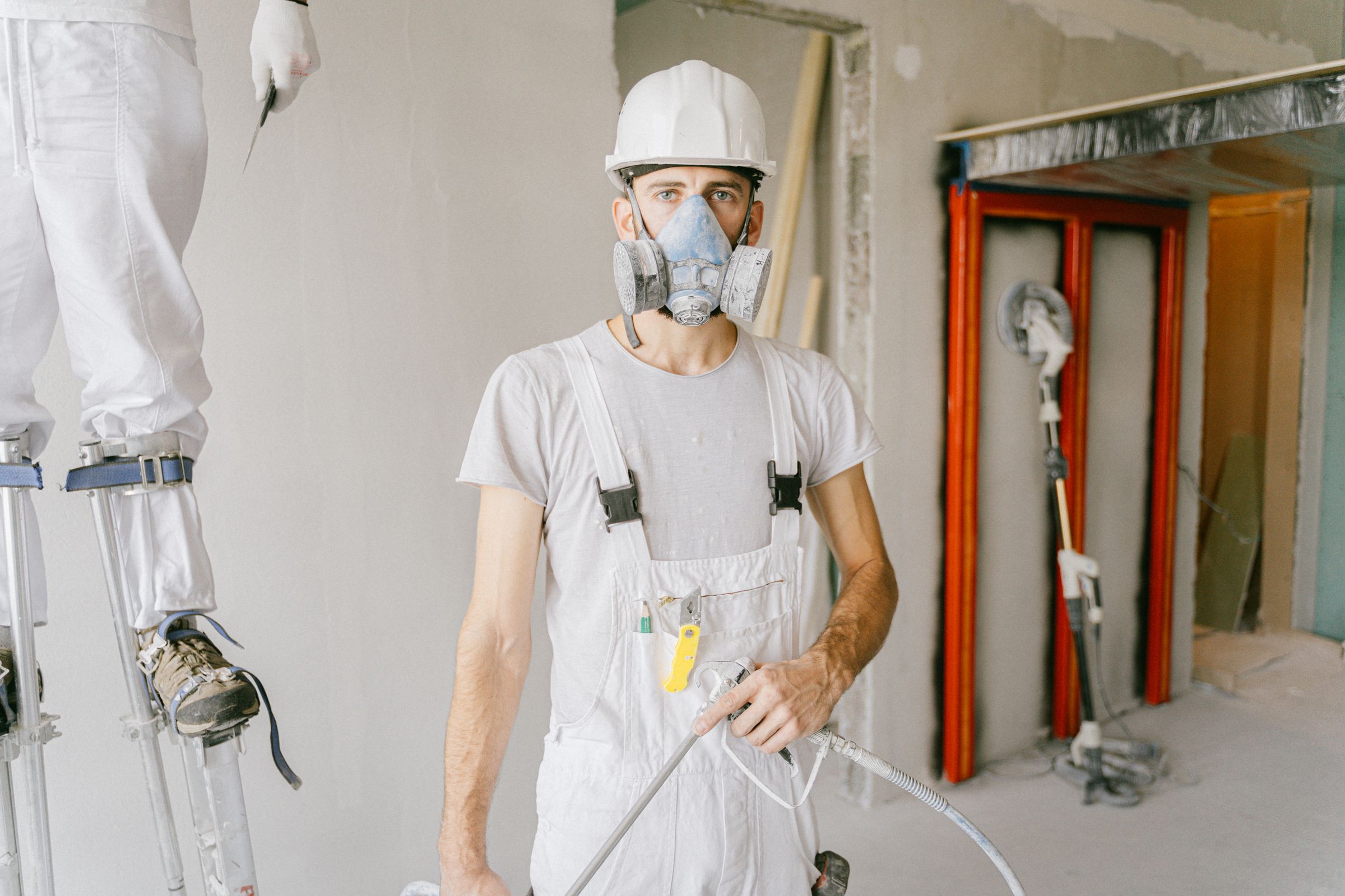 A professional painter in coveralls and respirator holding a paint spray gun on a jobsite