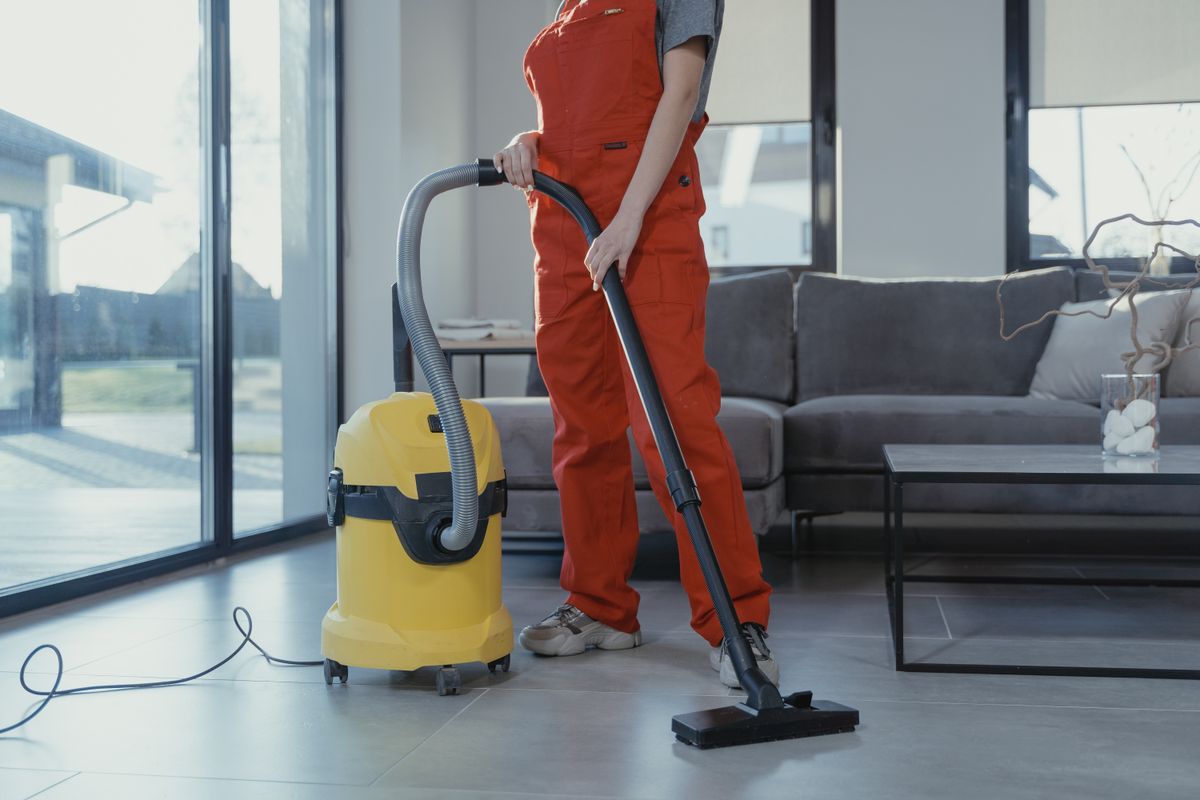 Cleaning professional in orange uniform vacuuming a bright living room with large windows during a recurring home cleaning service