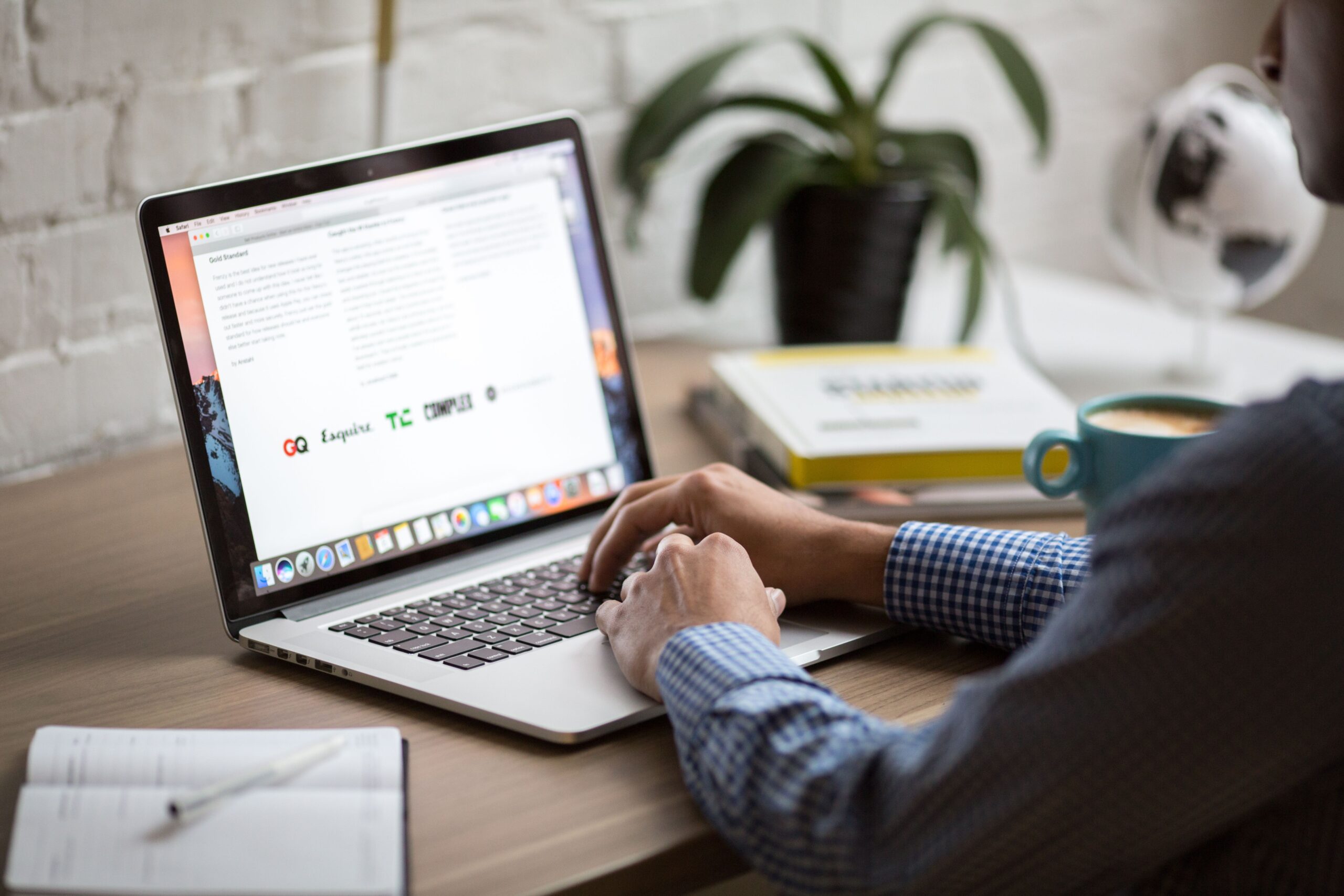 Person typing on MacBook at a desk with books, coffee, and a plant — writing content for a business website