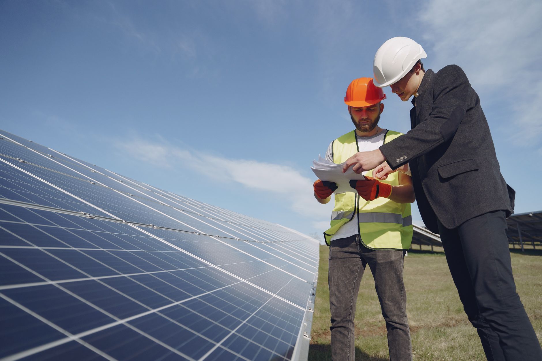 A solar installer in a hard hat reviewing an install proposal with a homeowner beside a ground-mounted panel array
