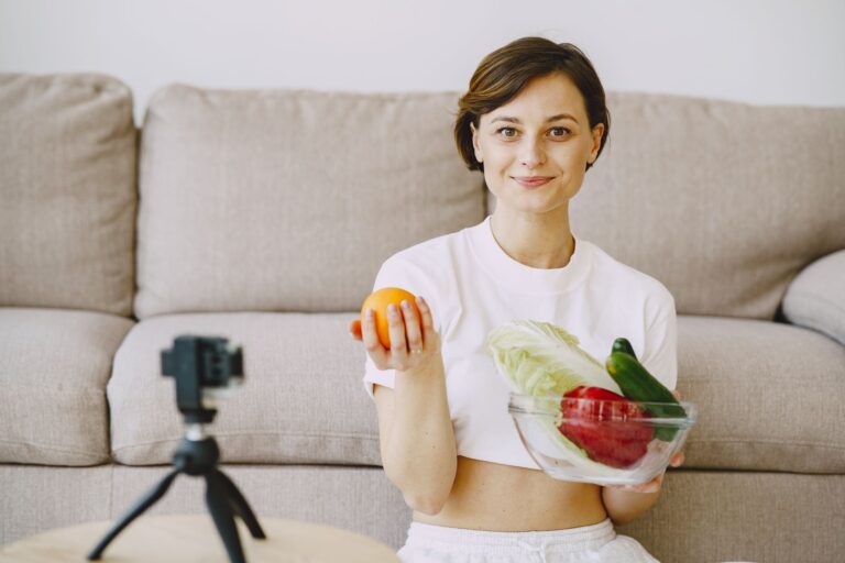 Optimistic female in white clothes sitting near sofa and holding bowl with fresh vegetables and oran