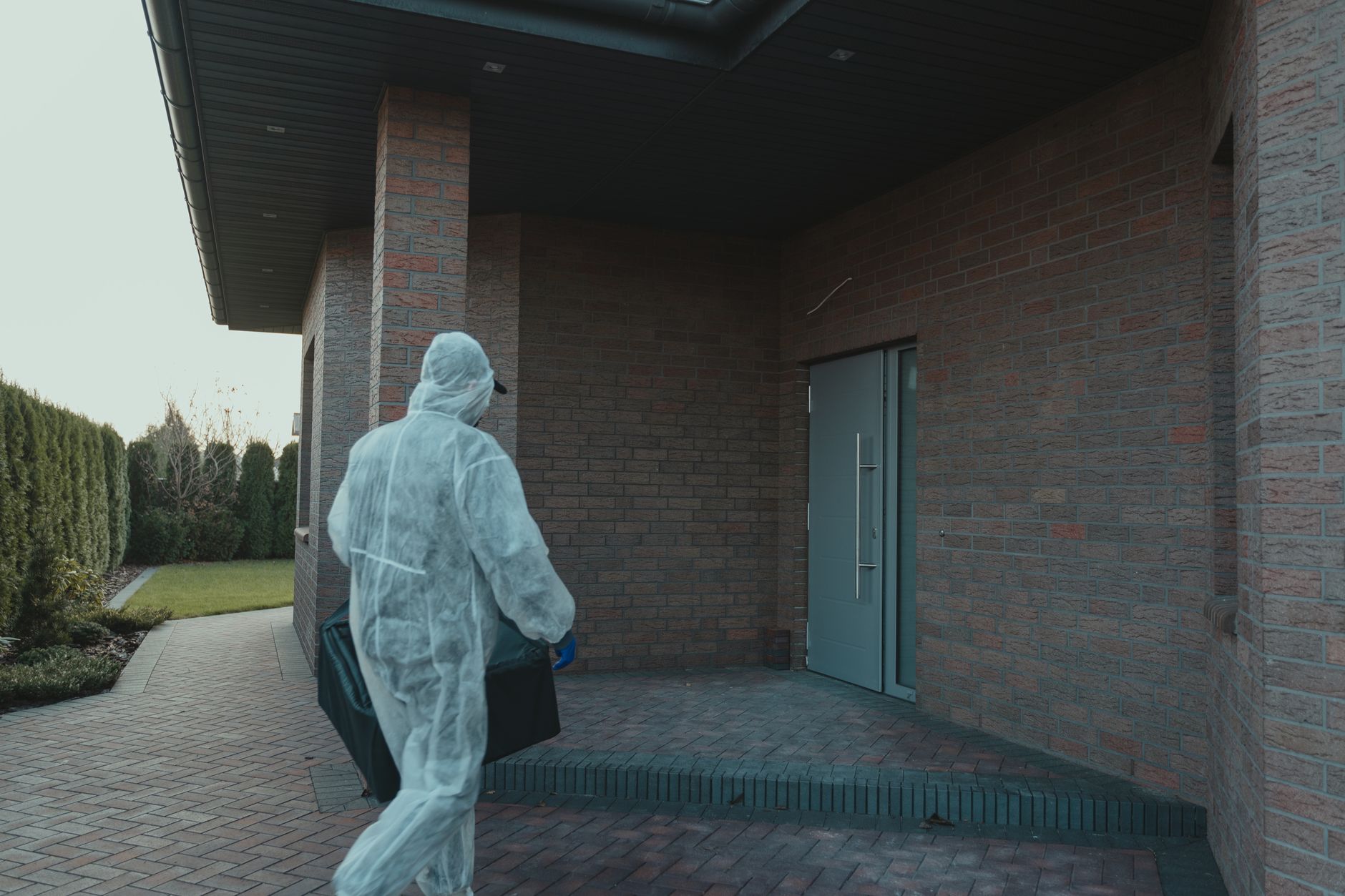 A person in PPE gear delivering a package to a contemporary brick house.
