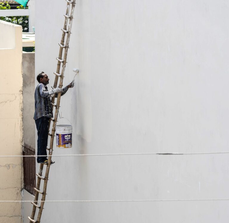 A painter on a ladder using a roller to paint a house exterior wall