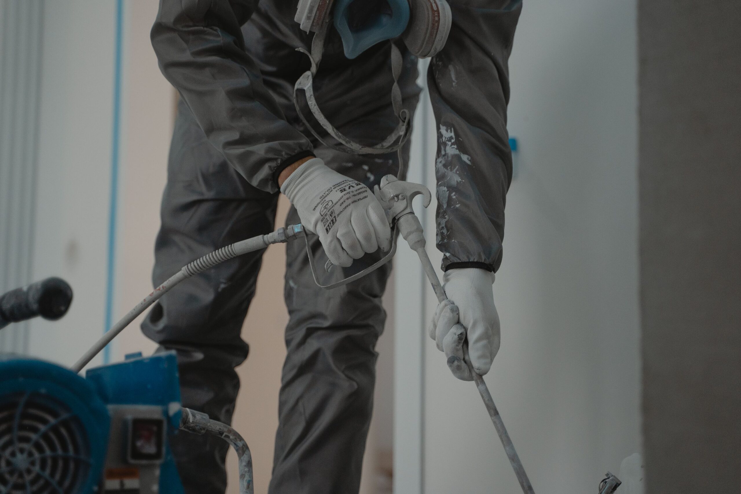 A worker in protective gear using a power sprayer to paint an interior space