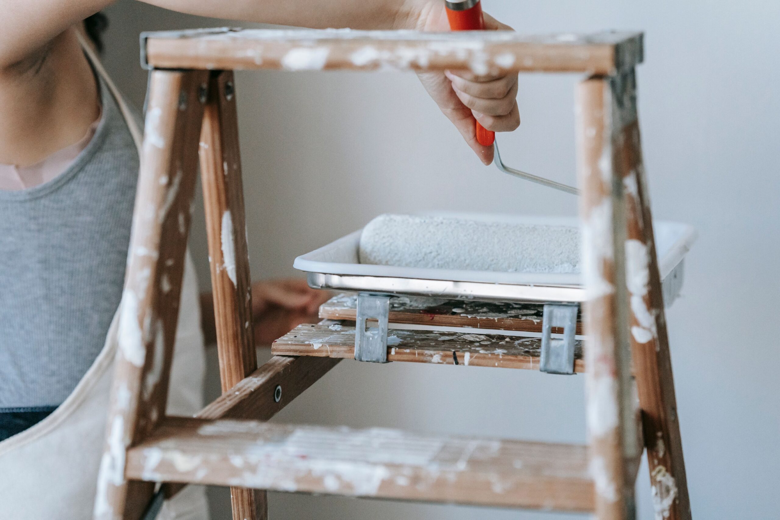 A woman using a roller and ladder to paint a room wall indoors