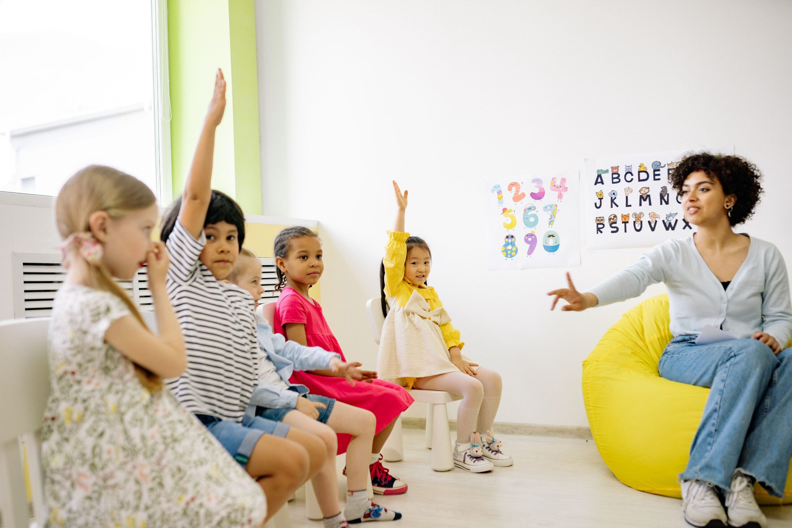 Preschool teacher engaging with young children raising their hands in a bright classroom