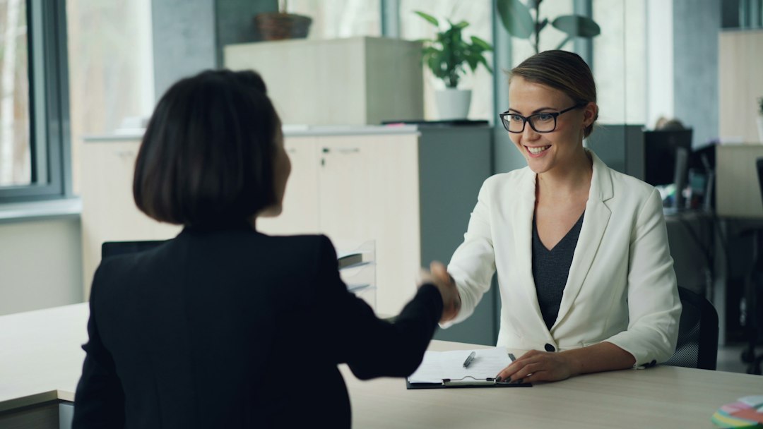 Two businesswomen shaking hands across a desk after closing a deal